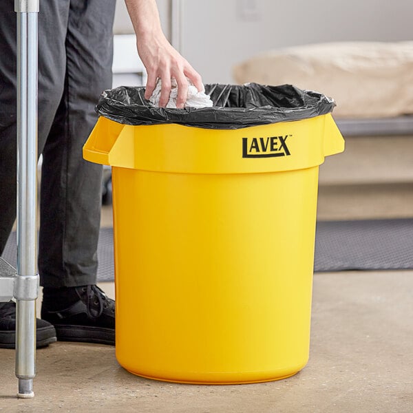 A person putting a black bag in a yellow Lavex 20 gallon trash can.