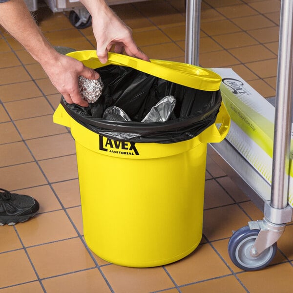 A hand placing foil in a yellow Lavex trash can with a lid.
