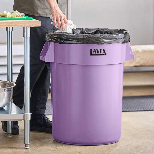 A man in black gloves putting a black plastic bag in a purple Lavex round commercial trash can.