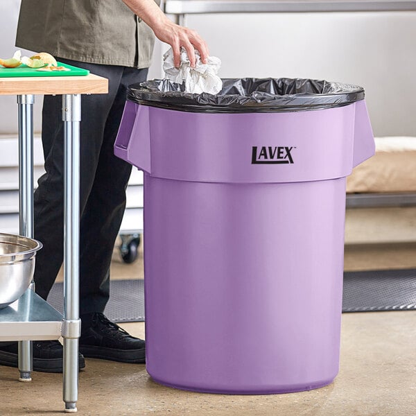 A man standing next to a Lavex purple round commercial trash can.