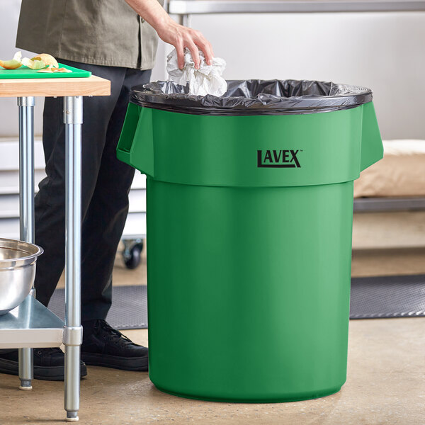 A man standing next to a Lavex green commercial trash can with a black plastic bag inside.