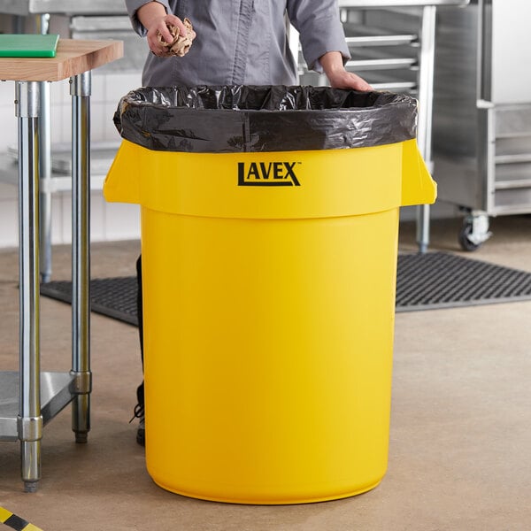 A woman standing in a school kitchen next to a yellow Lavex round commercial trash can.