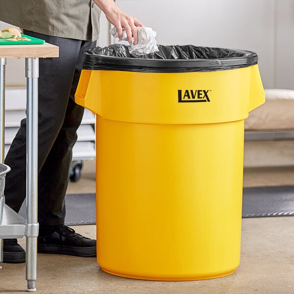 A man putting a plastic bag in a yellow Lavex 55 gallon trash can.