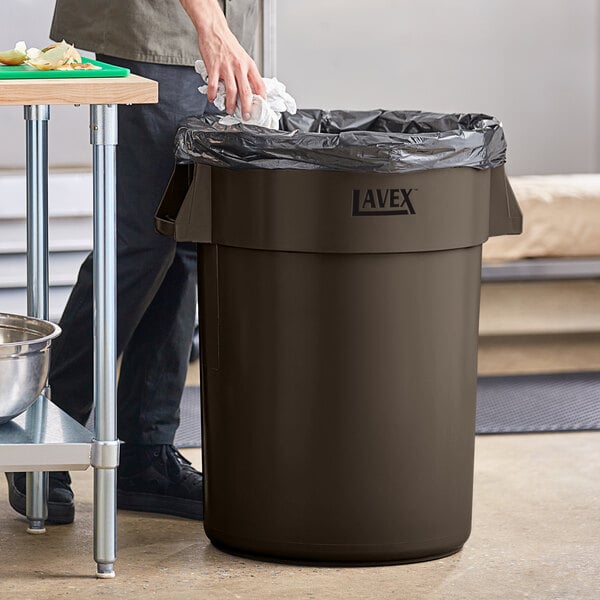 A man standing next to a Lavex brown round commercial trash can.