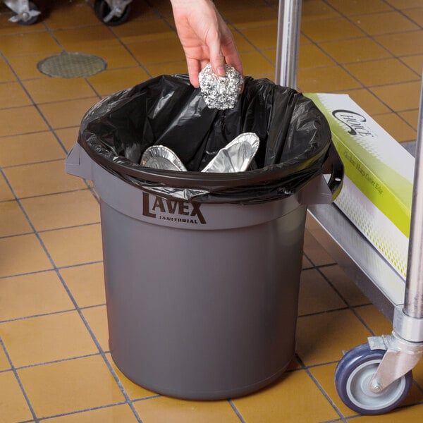 A person holding a Lavex gray round trash can with a silver lid.