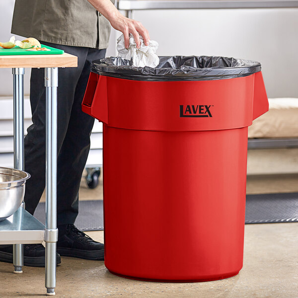 A man putting a black plastic bag in a red Lavex 55 gallon round trash can.