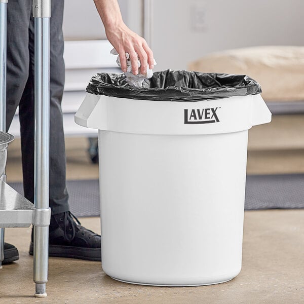 A person putting a black bag into a white Lavex round commercial trash can.