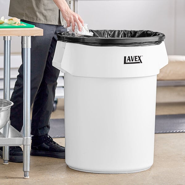 A person putting a plastic bag in a Lavex white round commercial trash can.