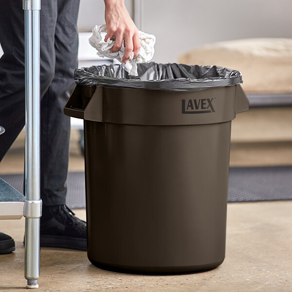 A man putting a plastic bag in a brown Lavex round commercial trash can.