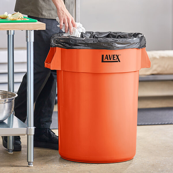 A man in black pants putting a black bag in an orange Lavex round commercial trash can.