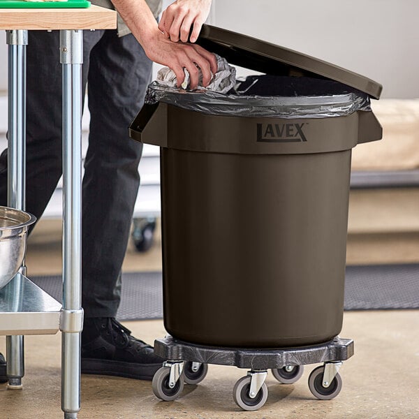 A man using a dolly to open a Lavex 20 gallon brown round commercial trash can with a lid to put a trash bag in.