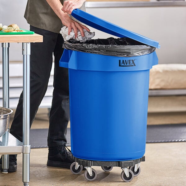 A man opening a blue Lavex 32 gallon commercial trash can in a school kitchen.