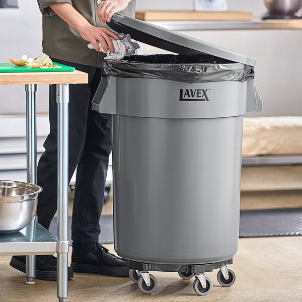 A man standing next to a Lavex 44 gallon gray commercial trash can.