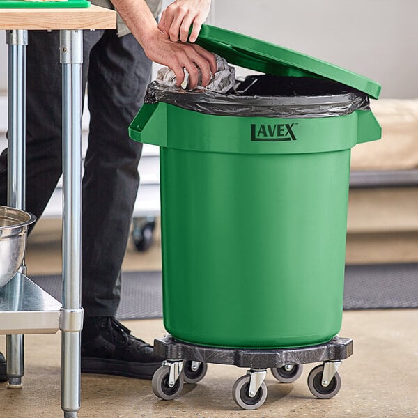 A man using a dolly to put a plastic bag in a Lavex green trash can.