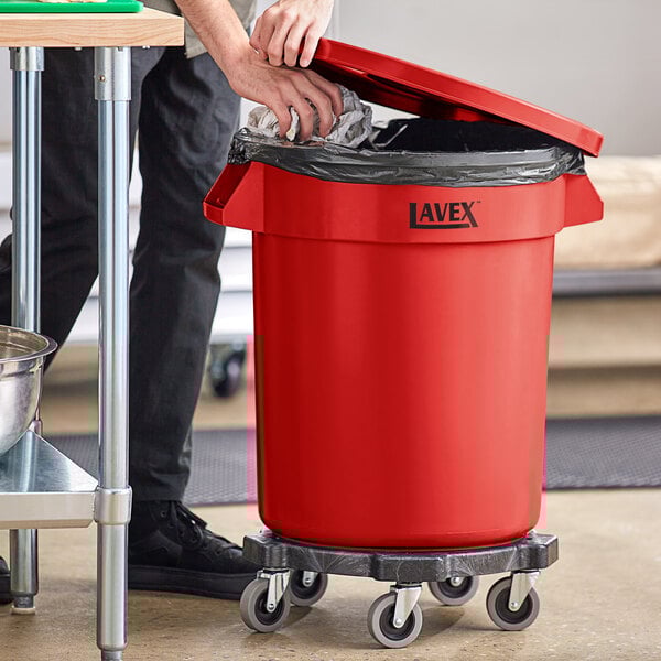A man using a dolly to open a red Lavex commercial trash can with a black bag inside.