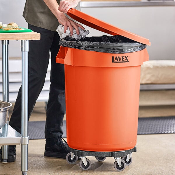 A man using a metal dolly to open an orange Lavex trash can in a school kitchen.