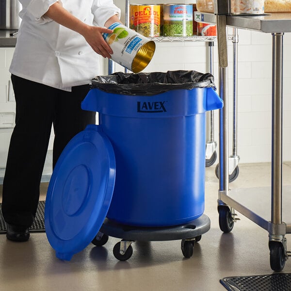 A woman pouring liquid into a Lavex blue round trash can in a school kitchen.