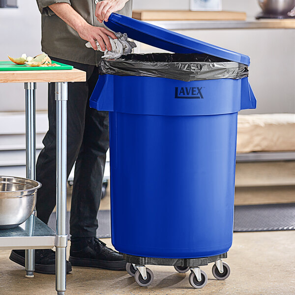 A man standing in a professional kitchen next to a blue Lavex commercial trash can.