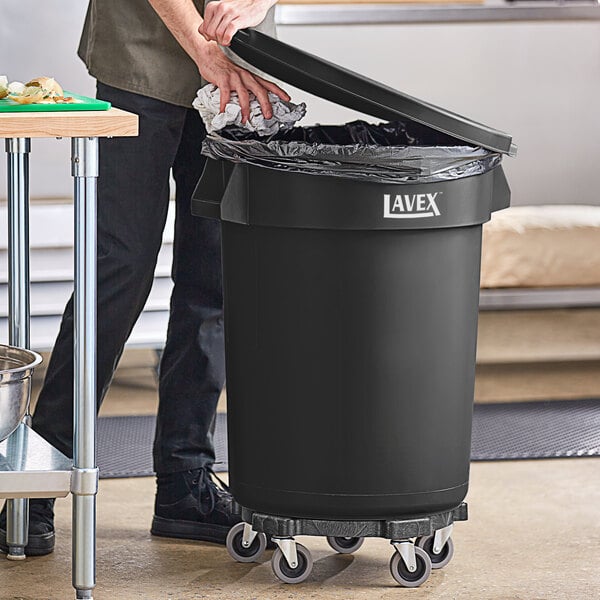 A man using a dolly to open a black Lavex commercial trash can to put a trash bag in.