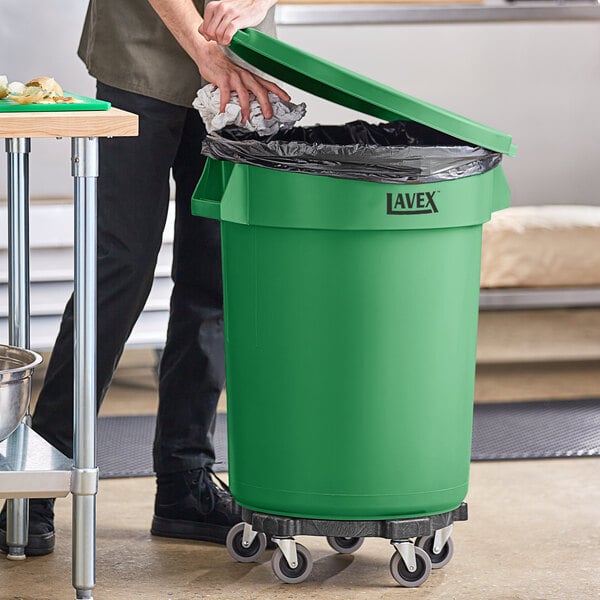 A man standing next to a Lavex green trash can with lid and dolly opening it to put in a plastic bag.