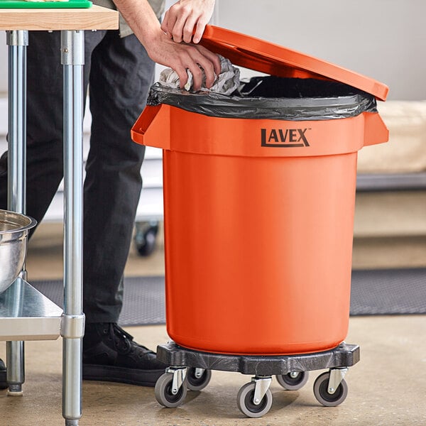 A man using the handle to open a Lavex orange trash can and put a bag of garbage in it.