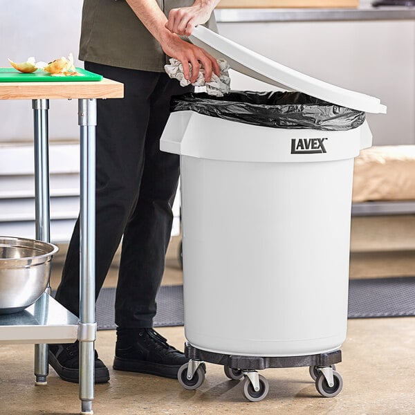 A man wearing black pants and shoes standing next to a white Lavex round commercial trash can with a black dolly lid.