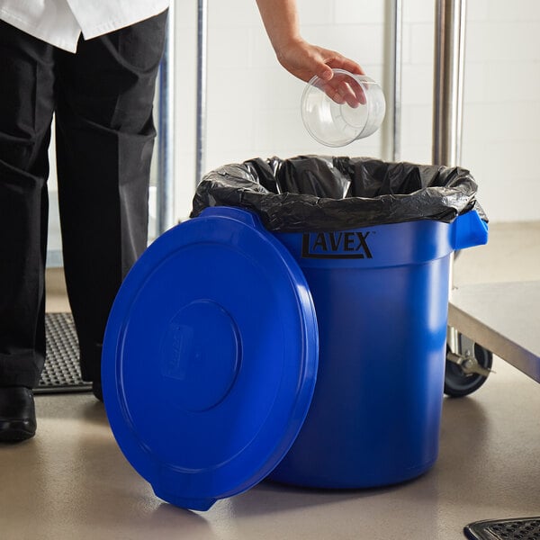 A woman putting a plastic cup into a blue Lavex trash can.