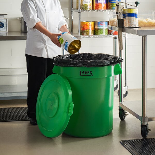 A woman in a kitchen pouring a can into a green trash can.