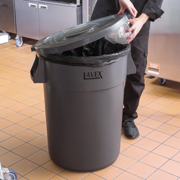 A person putting a plastic bag into a Lavex 44 gallon gray round commercial trash can.