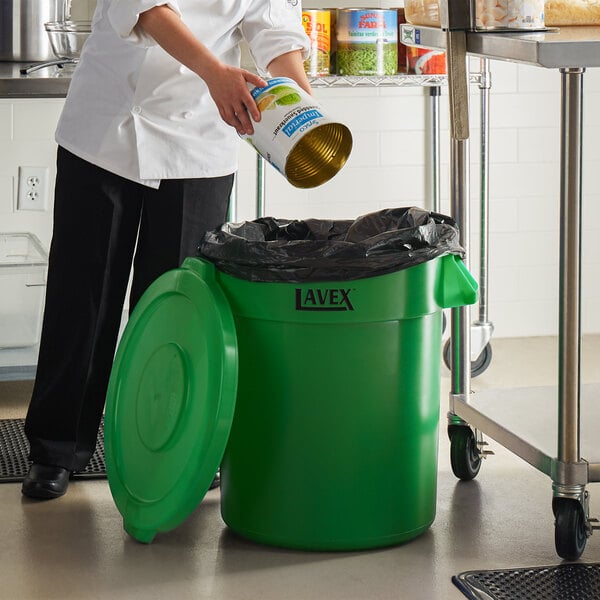 A woman in a white coat pouring food from a can into a green Lavex trash can.