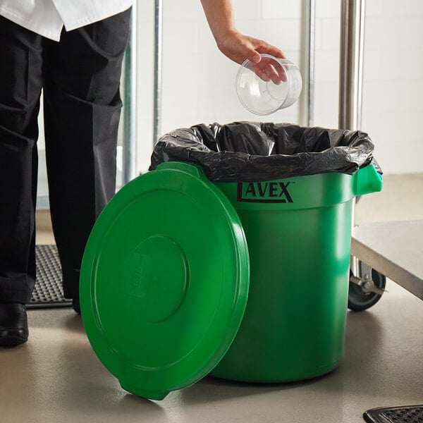 A woman putting a plastic cup into a green Lavex trash can.