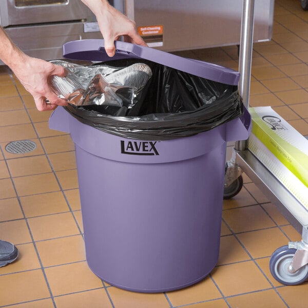 A hand putting foil in a Lavex purple round trash can with a lid.