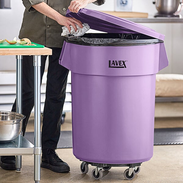 A woman standing in a professional kitchen is putting a black bag in a purple Lavex commercial trash can on a dolly.