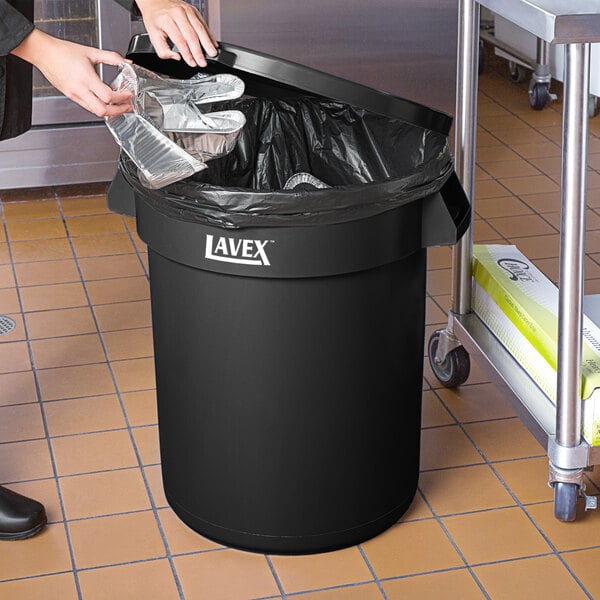 A person's hands holding a plastic bag and putting it into a Lavex 32 gallon round black trash can.
