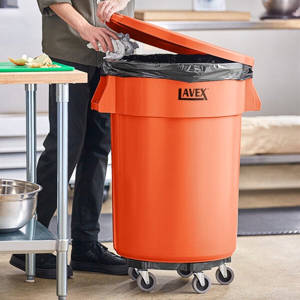 A man standing next to a large orange Lavex commercial trash can with a lid on it.