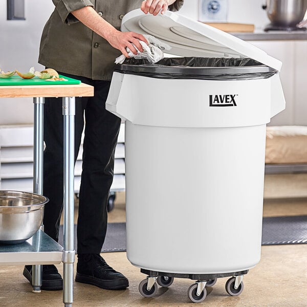 A woman standing next to a white Lavex commercial trash can with a black lid in a professional kitchen.