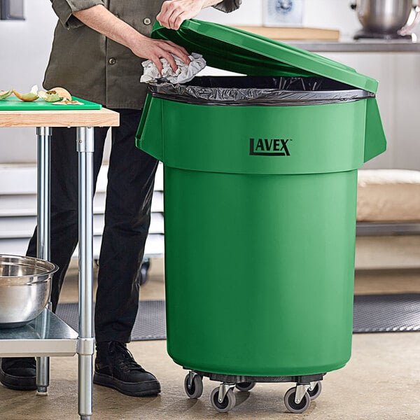 A woman in a professional kitchen standing next to a green Lavex trash can with a black lid.