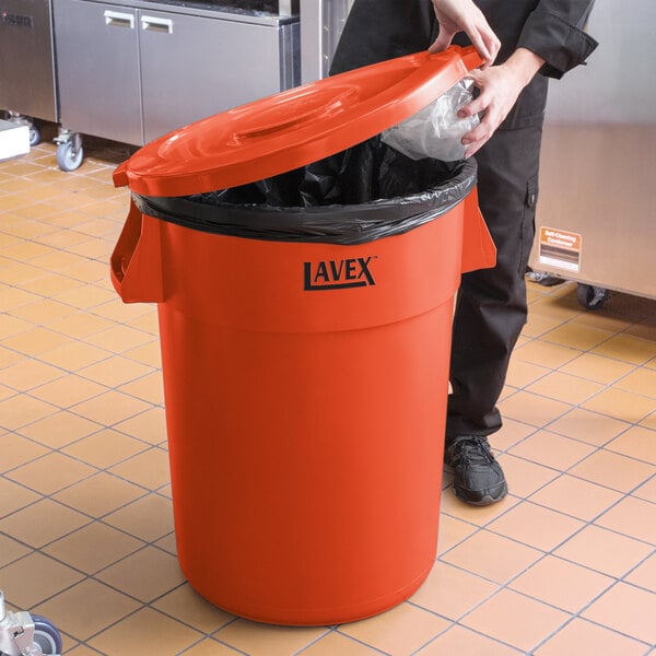 A person holding a plastic bag in a Lavex orange round trash can with a lid.