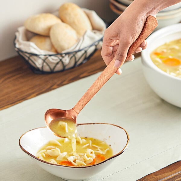 A person holding an American Metalcraft hammered bronze ladle over a bowl of soup.