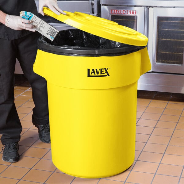 A man in a chef's uniform putting a carton in a yellow Lavex 55 gallon trash can.