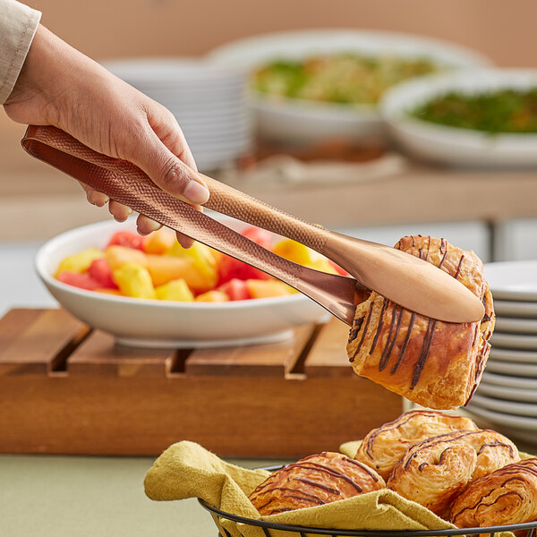 A person using American Metalcraft hammered bronze vintage tongs to pick up fruit from a bowl.