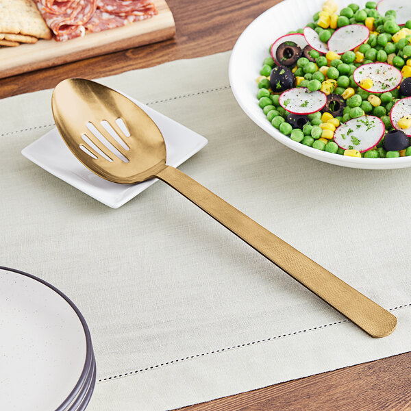 A gold slotted spoon on a plate of salad with a bowl of vegetables.