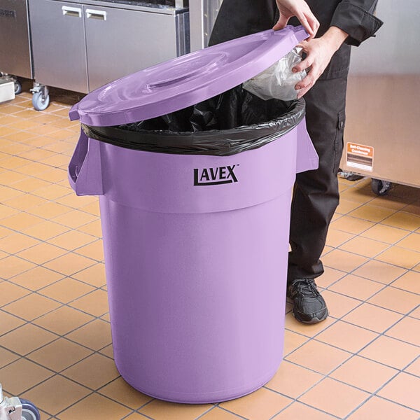 A person in black pants putting a plastic bag into a purple Lavex trash can with a lid.
