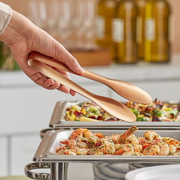 A hand holding American Metalcraft bronze tongs over a tray of food.