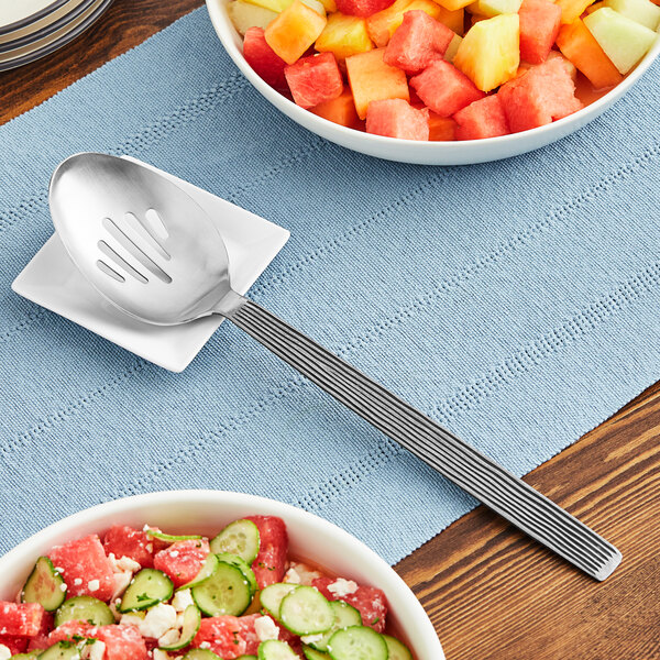 An American Metalcraft wavy stainless steel slotted spoon in a bowl of fruit salad.