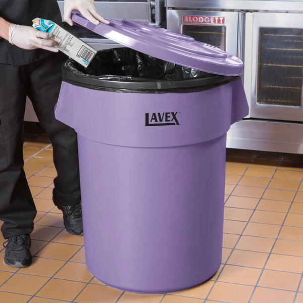 A man in a chef's uniform putting a package into a purple Lavex 55 gallon trash can.