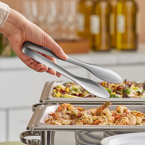 A person holding a pair of American Metalcraft stainless steel tongs over food on a buffet table.