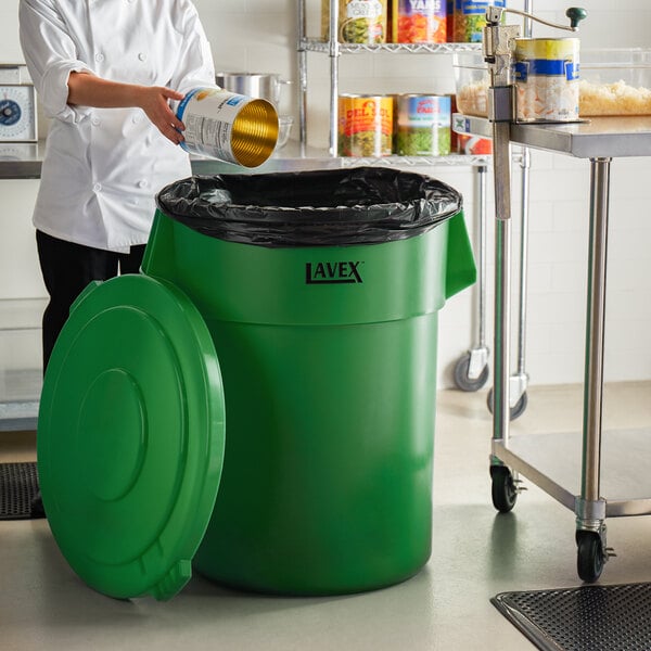 A woman pouring a can into a Lavex green trash can in a school kitchen.