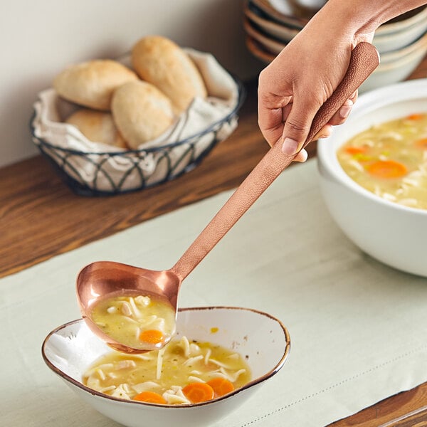 A person holding an American Metalcraft hammered bronze ladle over a bowl of soup.