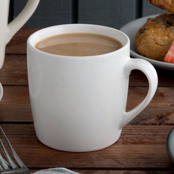 A close-up of a Tuxton Pearl White China Mug filled with brown liquid on a saucer with cookies.